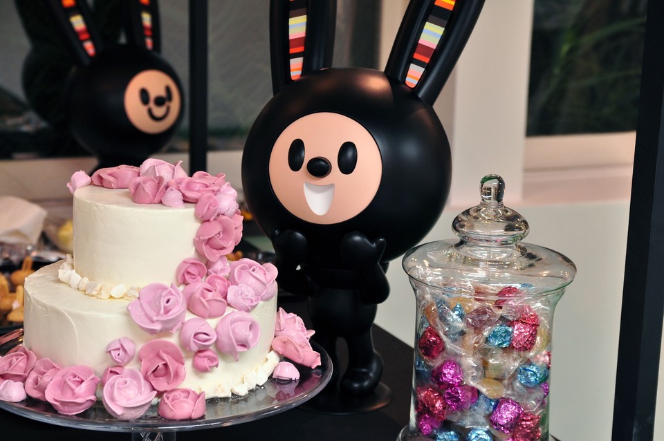 A black LUCKY BUNNY LUNNY sculpture with striped ears beside a pink flower-covered cake and a glass jar of colourful sweets.