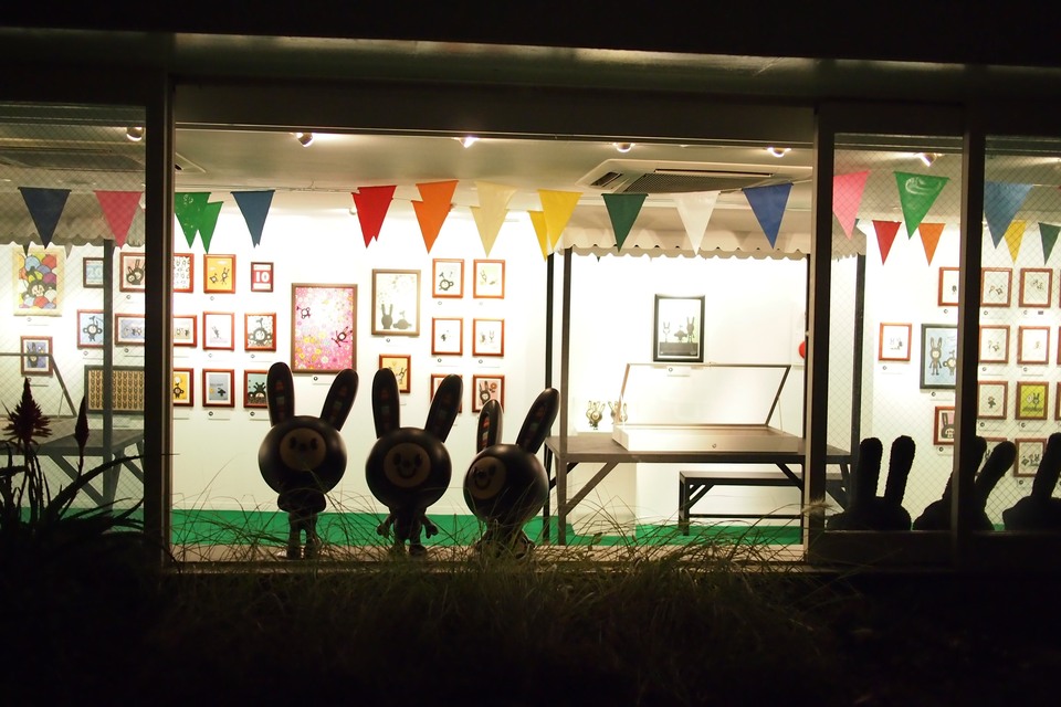 The gallery seen through its windows at night, with framed artworks across the walls, colourful bunting overhead and rabbit sculptures by the windows.